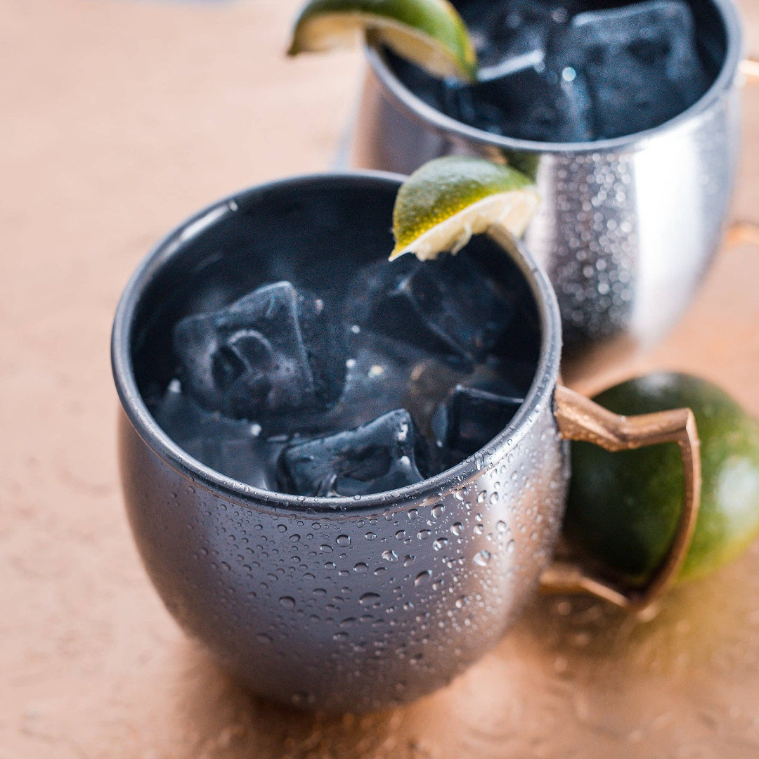Two dark gray ceramic mugs filled with iced drinks and garnished with lime wedges, set on a warm metallic surface with one mug in the foreground and another blurred in the background.