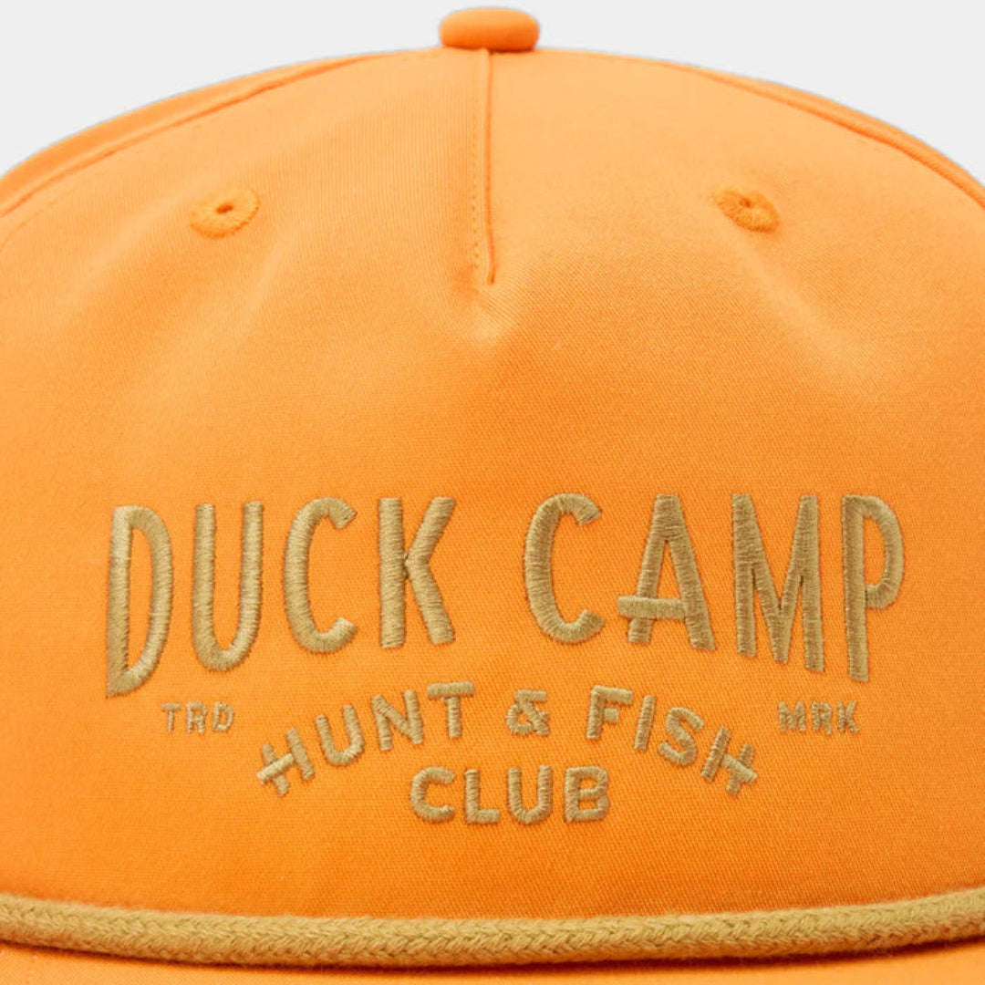 Close-up of an orange snapback cap with gold embroidered text reading 'DUCK CAMP' and smaller text 'HUNT & FISH CLUB' on the front panel.