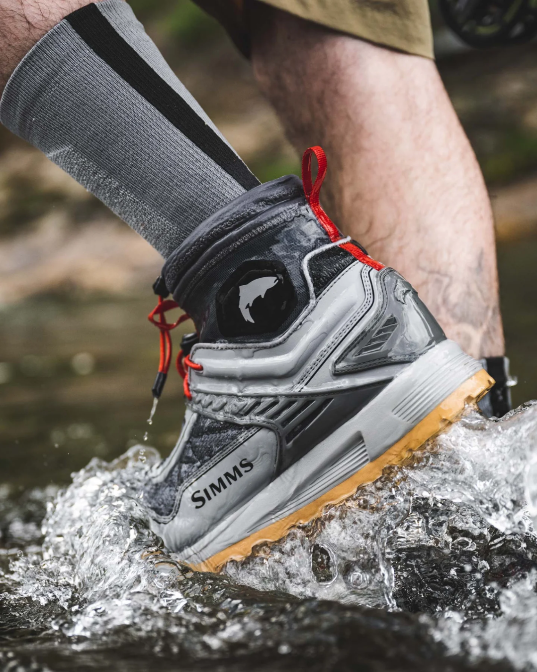Person wearing a gray and black Simms shoe with red laces, walking through water.