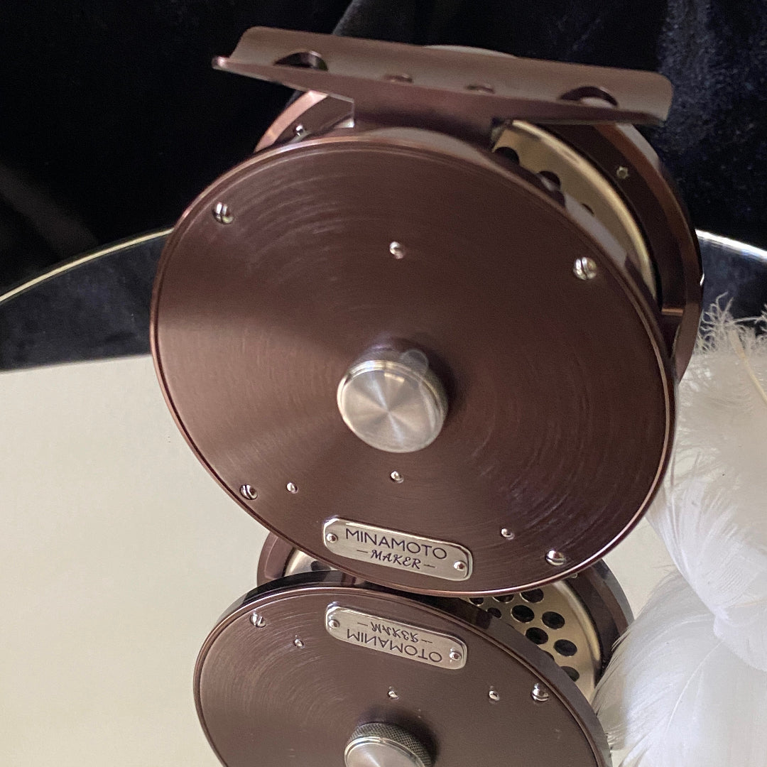 Close-up of two Minamoto M-6X fly fishing reels stacked vertically, showcasing their bronze-brown metal bodies, silver keyways, and engraved brand plates.