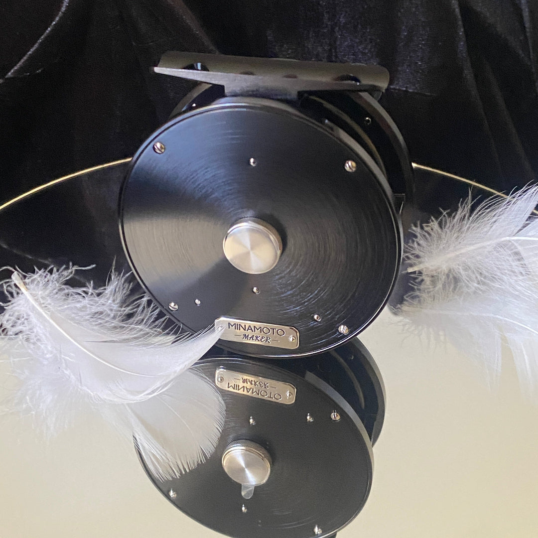 A black Minamoto-Marchi fly reel with a brushed metal finish, featuring a silver center knob and a silver brand plate. White feathers are tucked behind the reel, which is set against a black velvet background and reflected on a light surface below.