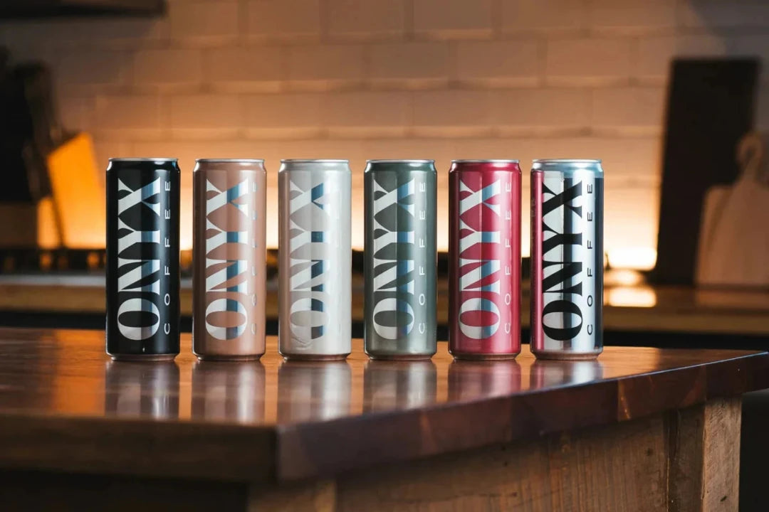 Six Onyx can beverages lined up on a wooden counter in a warm, dimly lit bar or kitchen setting.
