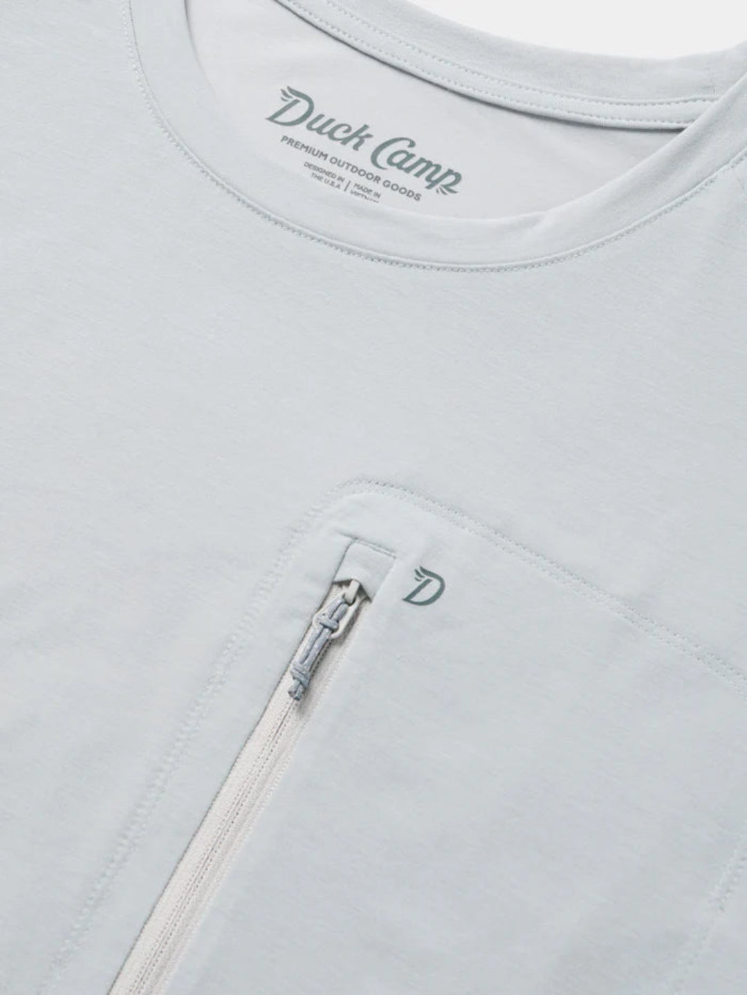 Close-up of a light gray or off-white zippered pocket on a matching crewneck shirt, with the Duck Camp brand name printed inside the collar and a small embroidered D logo on the pocket.