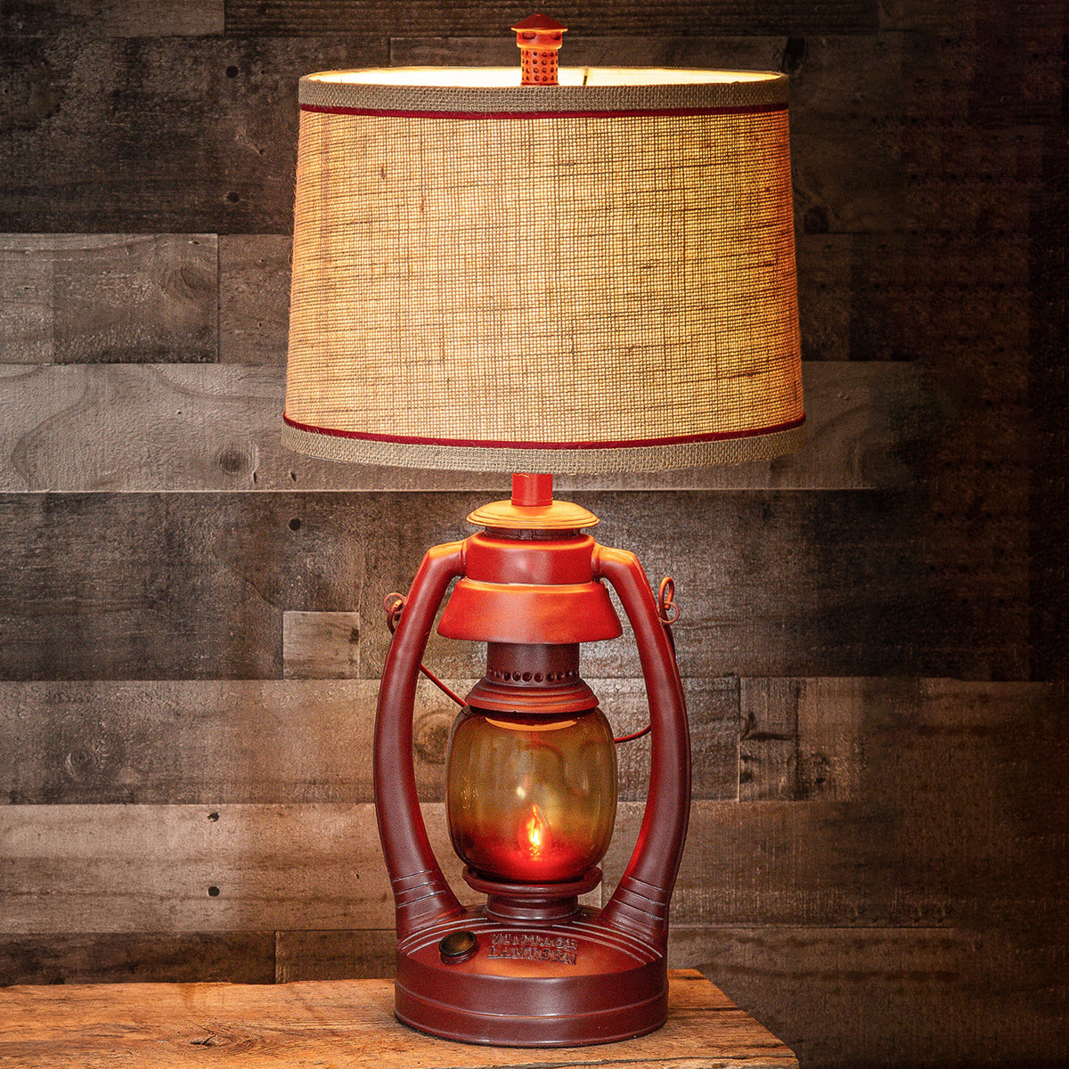 A rustic table lamp designed like a vintage lantern, featuring a warm orange-brown metal body, a glowing amber glass globe, and a large beige woven burlap shade with red trim, set against a dark wood plank background.