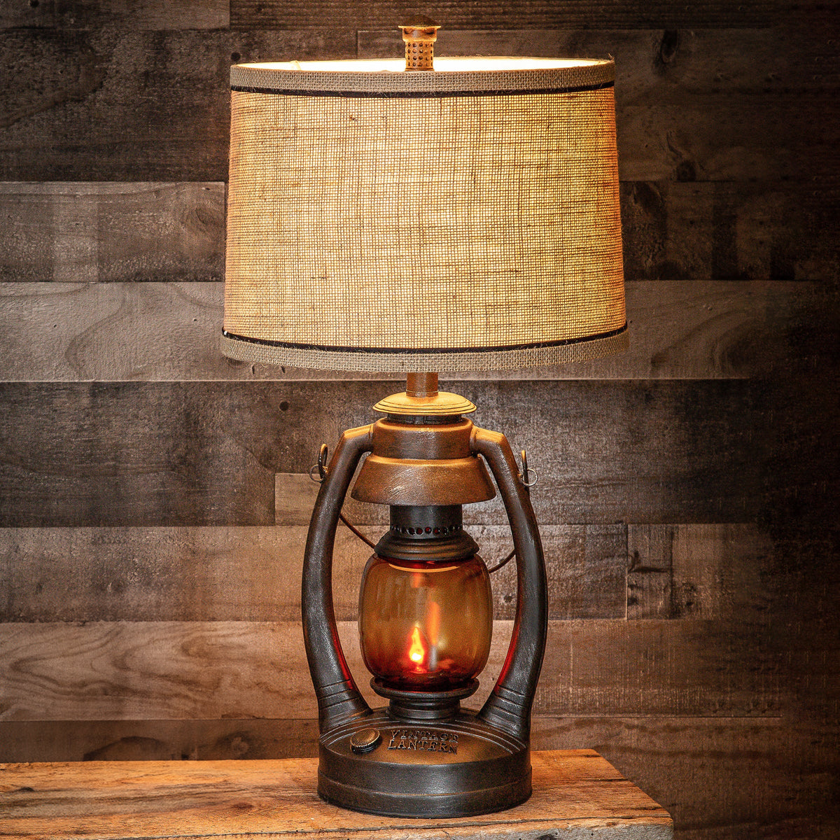 A rustic table lamp styled like a vintage oil lantern with a textured beige burlap drum shade and an amber glass lantern base, set against a dark wood plank wall.