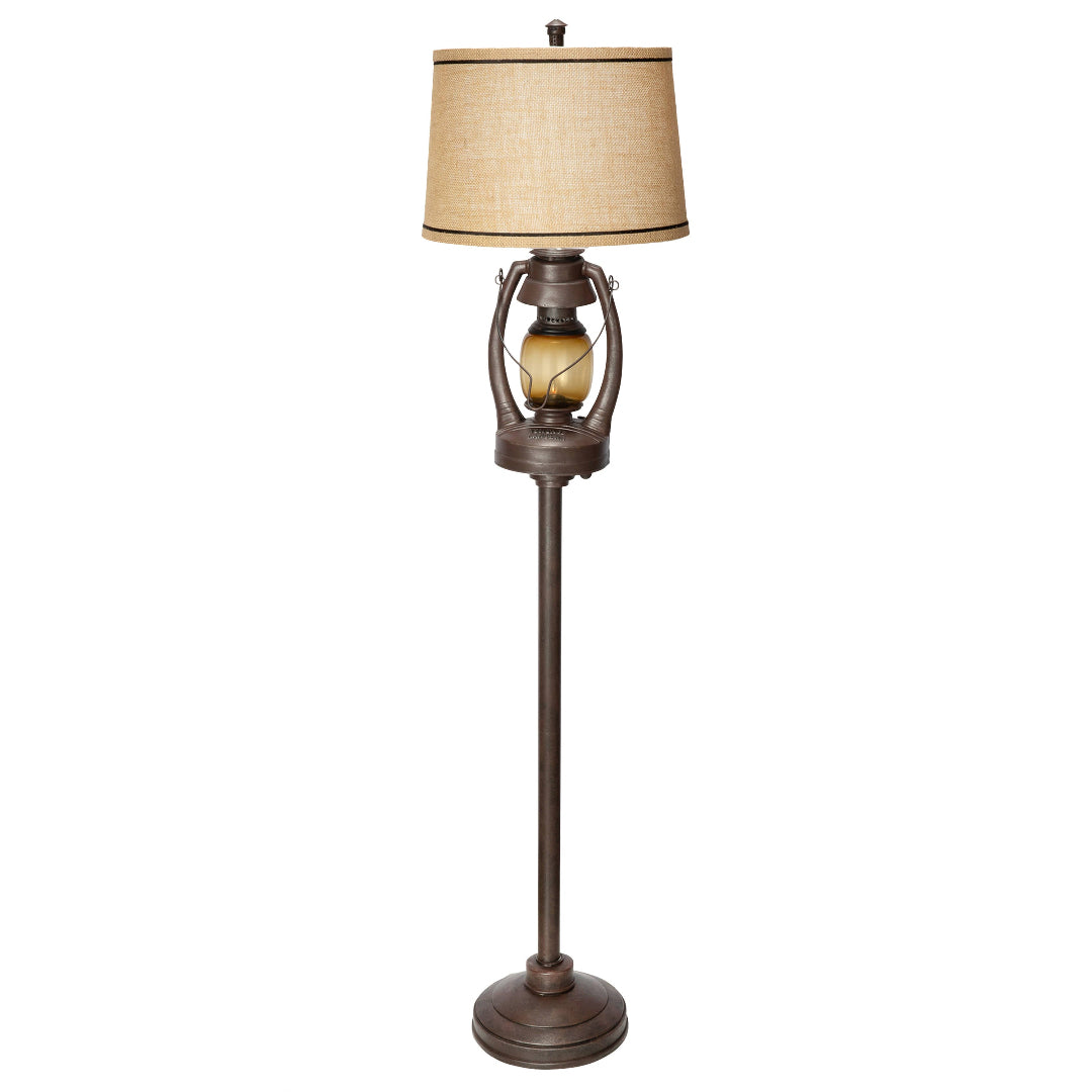 A rustic-style floor lamp with a tall dark bronze metal stand, a textured beige fabric drum shade, and a decorative amber glass bulb, shown against a plain white background.