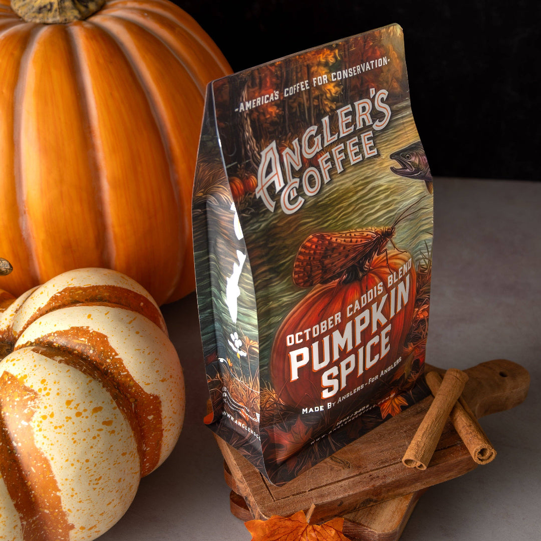 A bag of Angler's Coffee Pumpkin Spice coffee stands on a wooden board in a styled autumn scene with pumpkins and cinnamon sticks. The bag features a fall hunting and fishing theme with a large orange pumpkin graphic and the text 'October Caddis Fly Pumpkin Spice'.